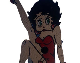 Puede incluir: Recorte de madera de una mujer de dibujos animados con vestido rojo, pelo negro y zapatos rojos. El recorte está pintado con colores negro, rojo y tono piel. La mujer está en una pose sentada con una pierna levantada. El recorte cuelga de una cuerda negra.