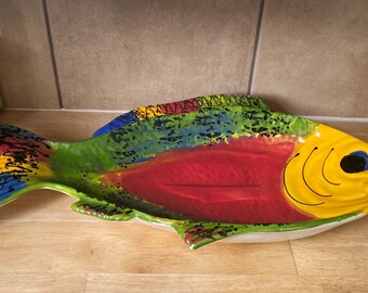 May include: A colorful ceramic fish-shaped serving platter. The platter features a yellow head, a red body, and a green and blue patterned back and tail. The fish-shaped dish is on a wooden surface.