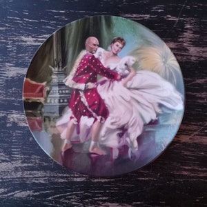 May include: A vintage porcelain plate featuring a painted scene of a couple dancing. The woman is wearing a white gown and the man is wearing a red and white patterned suit. The scene is set in a ballroom with a chandelier and other decorative elements.