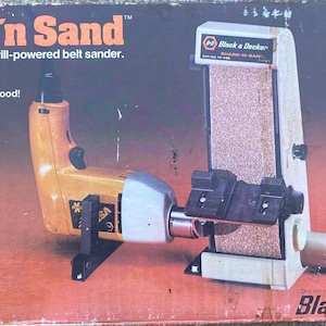 May include: A yellow and gray drill-powered belt sander with the text "Sharp'n Sand" and "Drill-powered belt sander."  The sander is attached to a white base with a black and gray belt sander. The box also has the text "Black & Decker" and "74-495".