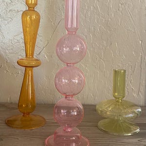 Op de afbeelding: Drie decoratieve glazen kandelaars in amber, roze en geel. De amberkleurige houder heeft een lang, slank ontwerp. De roze houder heeft een gestapeld bolontwerp. De gele houder heeft een brede, platte basis.