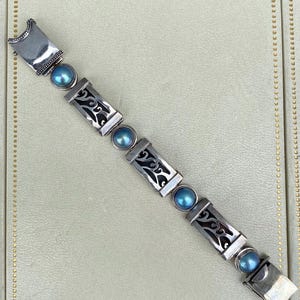 May include: A silver bracelet with a decorative design and blue pearls. The bracelet has a clasp closure.