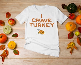 Könnte beinhalten: Weißes T-Shirt mit dem Text "I CRAVE TURKEY" in orangefarbenen Buchstaben. Unter dem Text befindet sich ein Cartoon-Bild eines gebratenen Truthahns. Das Shirt ist von Herbstblättern und Kürbissen umgeben.