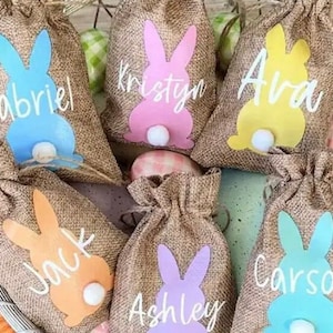 May include: Six burlap Easter bunny gift bags with personalised names: Gabriel, Kristyn, Ava, Jack, Ashley, and Carson. Each bag features a bunny silhouette with a fluffy tail.