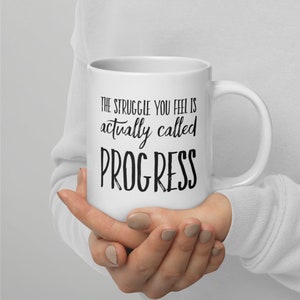 May include: White ceramic coffee mug with black text that reads "The struggle you feel is actually called PROGRESS".