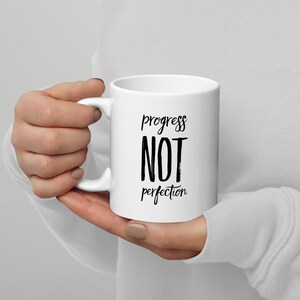 以下が含まれることがあります： 白地に黒文字で「progress not perfection」と書かれたセラミック製のマグカップです。