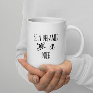 Op de afbeelding: Witte keramische koffiemok met zwarte tekst die "Be a dreamer and a doer" luidt.