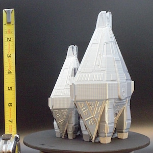 Puede incluir: Dos modelos plateados de aspecto futurista con diseños geométricos detallados. El modelo más grande mide aproximadamente 13 cm de altura. Los modelos se exhiben sobre una superficie oscura, con una cinta métrica amarilla en el fondo.