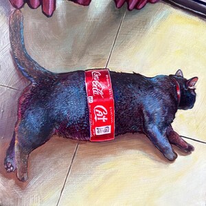 May include: A black cat with a red Coca-Cola can strapped around its middle. The cat is lying on a light brown surface.