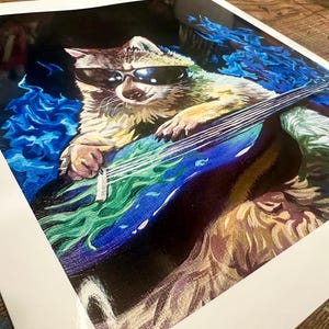 Pu&ograve; includere: Un procione con occhiali da sole suona una chitarra elettrica blu con accenti verdi e bianchi. Il procione &egrave; circondato da fiamme blu.
