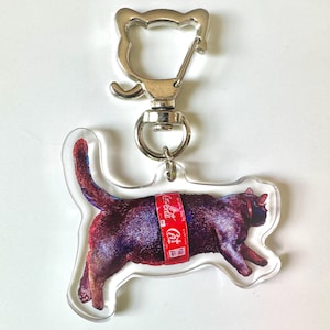 May include: A clear acrylic keychain featuring a black cat design with a red Coca-Cola band around its middle. The keychain has a silver-colored clasp and ring for attaching to keys or bags.