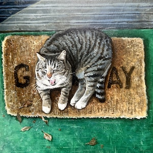 May include: A tabby cat resting on a doormat that reads "GAY". The cat is gray and white with black stripes. The doormat is brown and rectangular, and the background is green.