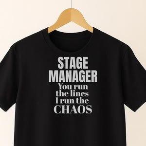 Pode incluir: T-shirt preto com o texto "STAGE MANAGER You run the lines I run the CHAOS" em letras brancas. A camisa está pendurada em um cabideiro de madeira.