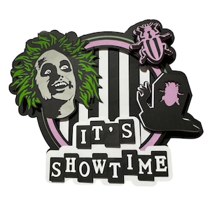 Puede incluir: Una decoración de papel recortado en capas que presenta un personaje con cabello verde alborotado, un fondo de rayas blancas y negras, un escarabajo morado, una lápida negra con la silueta de un escarabajo rosa y el texto IT'S SHOWTIME.