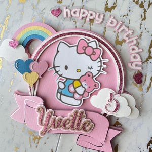 May include: A pink and white birthday cake topper featuring Hello Kitty holding a teddy bear. The topper includes a rainbow, clouds, balloons, the name "Yvette", and the number "5". The words "happy birthday" are also included.