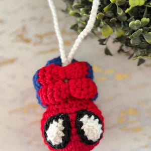 Może przedstawiać: Ręcznie robiona ozdoba na szydełku w kształcie Spider-Mana. Ozdoba jest gł&oacute;wnie czerwona z niebieskimi akcentami i ma czarno-białe oczy. Do zawieszenia przymocowany jest biały sznurek. Do użytku dekoracyjnego.