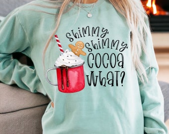 May include: A light blue sweatshirt with a graphic of a gingerbread man on top of a mug of hot cocoa with whipped cream and a red and white striped straw. The text on the sweatshirt reads "Shimmy Shimmy Cocoa What?"