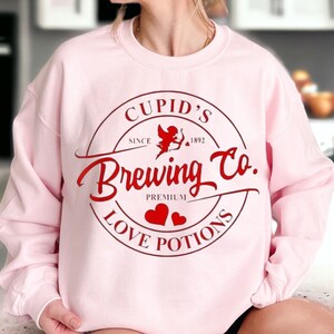 May include: Pink sweatshirt with a red graphic design that says "Cupid's Brewing Co. Since 1892 Premium Love Potions". The graphic is a circle with a red cupid inside and two red hearts below.