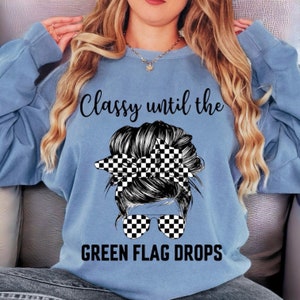 May include: A light blue sweatshirt with the text "Classy until the green flag drops" and a black and white checkered graphic of a woman's hair in a bun with sunglasses.