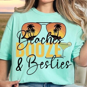 May include: A turquoise t-shirt with a graphic design of sunglasses with palm trees and a sunset. The text "Beaches Booze & Besties" is printed in yellow and black.