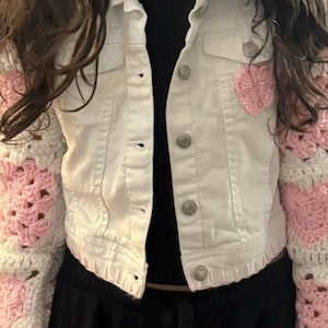 Puede incluir: Chaqueta vaquera blanca con detalle de corazón de ganchillo rosa en el pecho. Las mangas son de ganchillo blanco y rosa. La chaqueta tiene botones plateados y dos bolsillos en el pecho. Se ve una camiseta negra debajo.