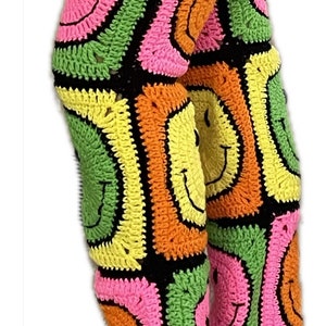Puede incluir: Un par de pantalones de crochet coloridos con un patrón de cara sonriente. Los pantalones están hechos con hilo verde, rosa, amarillo, naranja y negro.