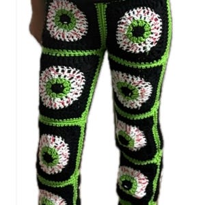 May include: Crocheted pants featuring a repeating eye design. The pants are made of black squares with green borders, each containing a white, red, and green eye motif. The handmade pants are a unique fashion statement.