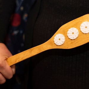 Puede incluir: Una paleta de madera con tres pomos redondos blancos texturizados unidos a la superficie. La paleta está sostenida por una mano.