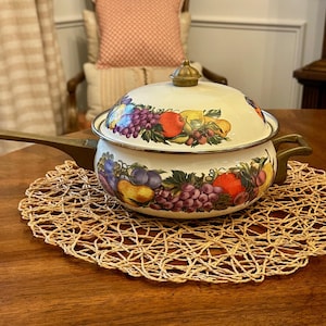 May include: A white enamel pot with a lid, featuring a colorful fruit design of grapes, apples, and pears. The pot has a long handle and a small handle on the side. It sits on a round, woven placemat.