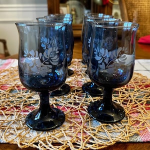May include: Set of four dark blue stemmed glasses with etched floral designs. The glasses have a tulip shape and sit on a woven placemat. The glasses are on a wooden table.