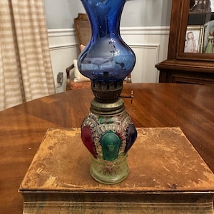 May include: An antique oil lamp with a cobalt blue glass top and a decorative base featuring red, green, and blue accents. The lamp sits atop a vintage, leather-bound book, showcasing a classic design.