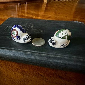 May include: Two small, hand-painted ceramic snail figurines with a crackled white glaze. The left snail features blue and brown floral patterns, while the right snail has green leaf designs and the text MEX. A silver coin sits between them.