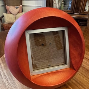 May include: A round, red wooden photo frame with a silver-colored frame insert. The frame has a circular design with a flat base and a recessed area to hold a photograph. The frame is sitting on a wooden surface.