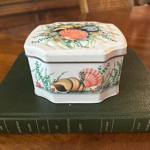 May include: A white octagonal metal tin featuring a decorative print of various seashells, including conch and scallop shells, interspersed with green seaweed. The lid and sides are adorned with these marine elements in shades of orange, tan, and brown.