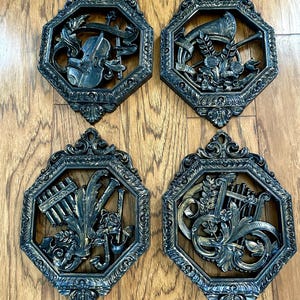 May include: Four vintage wall plaques with musical instrument designs. Each plaque features a different instrument: a violin, a trumpet, a pan flute, and a harp. The plaques are dark bronze with ornate, octagonal frames.