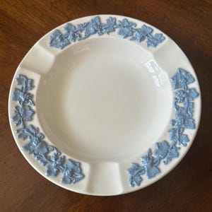 May include: A round, off-white ceramic ashtray featuring a raised, light blue relief pattern of grapevines and leaves encircling the rim. The design includes three evenly spaced notches along the edge for resting items, with a smooth, glossy finish.