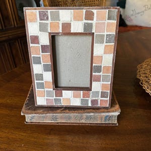 May include: A decorative photo frame with a mosaic tile design in shades of brown, beige, and gray. The frame is rectangular with a brown border and sits atop a vintage book. The frame is designed to hold a photograph.