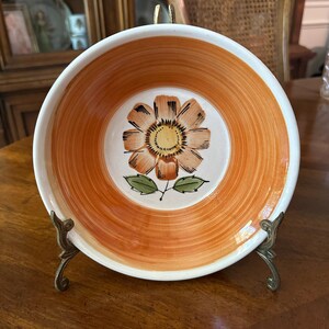 May include: A round ceramic bowl featuring a hand-painted orange brushstroke pattern on the interior walls and a central illustration of a single flower with light orange petals, a yellow center, and two green leaves on a white background.