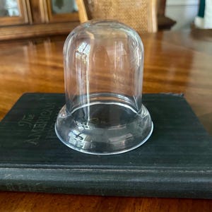May include: A clear, dome-shaped glass cloche with a smooth, rounded top and a flared base. The cloche is positioned on top of a dark, rectangular book with the visible text "The AMERICAN" printed in gold lettering on the front cover.