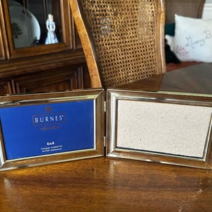 May include: A gold-toned, hinged photo frame, open to display two 6x4 inch photos. The frame has a blue backing on one side with the brand name "BURNES" and text. Lacquer coated for tarnish resistance.