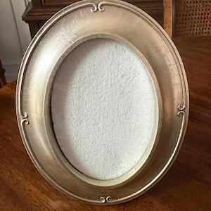 May include: Oval-shaped, antique-style picture frame with a metallic gold finish. The frame has decorative flourishes at the top and bottom. The interior is white, and the frame is sitting on a wooden surface.