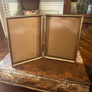 May include: A hinged, gold-toned metal picture frame with two rectangular openings. The frame is set on a distressed, antique book with peeling paint. The background includes a wooden cabinet and a chair.