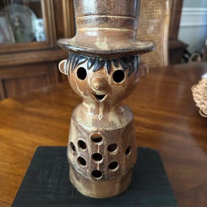 May include: A brown ceramic figurine shaped like a person wearing a top hat. The figure features a smooth, glazed finish with dark brown speckles, two circular eye cutouts, a small nose, a smiling mouth, and a series of circular holes on the torso.