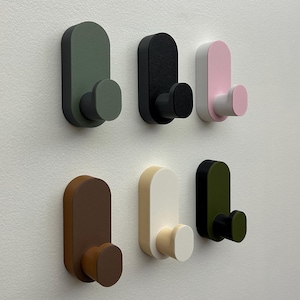 May include: Six wall hooks in various colors: olive green, black, pink and white, brown, cream, and dark green with black. Each hook has a rounded rectangular backplate and a circular hook. The hooks are mounted on a white wall.