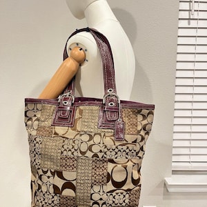 Puede incluir: Un bolso tote grande con un diseño de patchwork de varios paneles de tela con monograma en tonos marrón y beige. El bolso incluye ribetes de charol color burdeos oscuro, dos correas de hombro a juego, hebillas de metal plateado y una etiqueta de cuero.