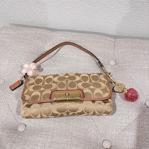 Puede incluir: Un bolso de mano con estampado de monograma en color tostado y marrón, con una correa de cuero, un cierre de metal dorado, un dije de flor rosa claro en el lateral y un dije esférico rojo brillante colgado de una anilla de metal dorado.