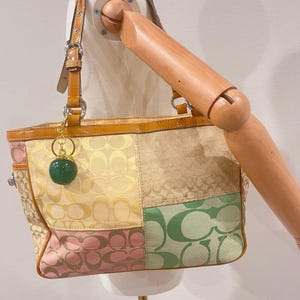 Könnte beinhalten: Eine Patchwork-Handtasche mit dem charakteristischen Coach-C-Logo-Muster auf verschiedenen Stoffbahnen in Beige, Hellgelb, Rosa und Grün. Die Tasche verfügt über beigefarbene Lederbesätze, silberfarbene Beschläge und einen runden, dunkelgrünen Glitzeranhänger.