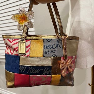 Puede incluir: Un bolso tote de Coach con un diseño de patchwork de varias texturas y colores, incluyendo paneles de oro metálico, rojo, azul y rosa. El bolso muestra el texto Coach est. 1941 New York, un colgante de flor y un lazo brillante con pedrería.