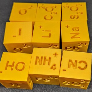 May include: A set of 10 yellow blocks with chemical formulas and symbols printed on them. The formulas include H₂O, NH₄, Fe, Li, Na, S₂O, CO₂, Pb, and O.