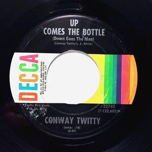 May include: A black vinyl record with a white label. The label has the text "Decca" in a large, colorful font. The record is titled "Up Comes The Bottle (Down Goes The Man)" by Conway Twitty. The record number is 32742 (7-122,605).